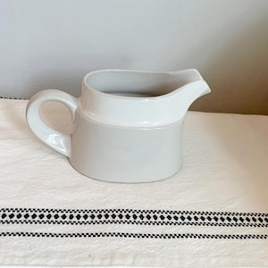 May include: A two-tone ceramic pitcher with a handle and spout. The top portion is white, and the bottom is a light gray. The pitcher is sitting on a white cloth with a black and white patterned border. The pitcher is designed for serving.