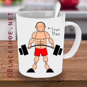 May include: White ceramic mug with a cartoon illustration of a muscular person lifting a barbell. The text "Iron Man" is written on the mug.