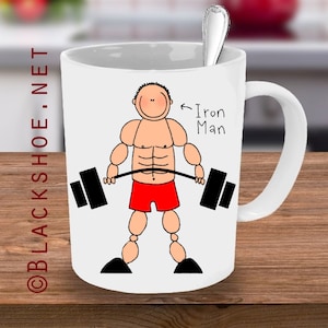 May include: White ceramic mug with a cartoon illustration of a muscular person lifting a barbell. The text "Iron Man" is written next to the person.