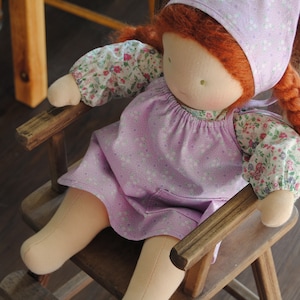 May include: A handmade doll with red hair, wearing a pink dress with white floral print and a pink bonnet, sitting in a wooden rocking chair.