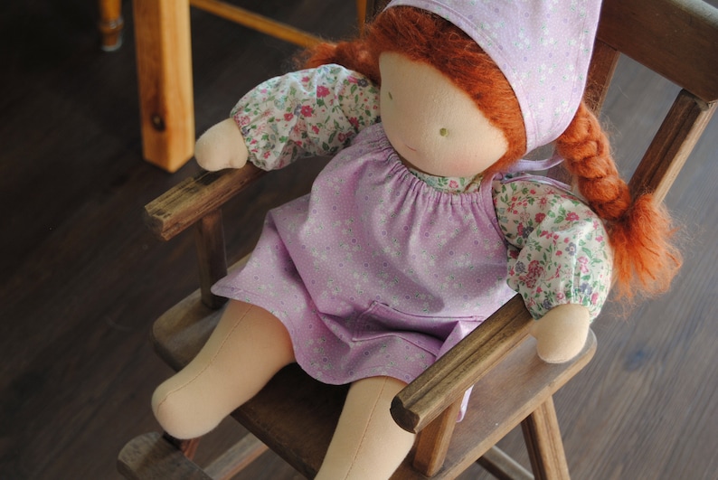 May include: A handmade doll with red hair, wearing a pink dress with white floral print and a pink bonnet, sitting in a wooden rocking chair.