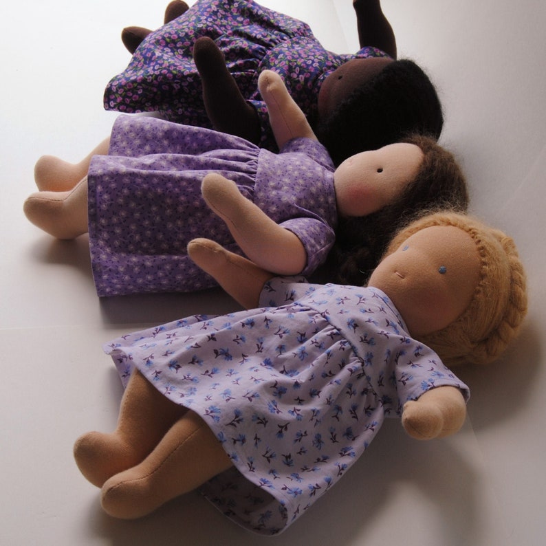 May include: Three handmade cloth dolls with purple and white floral print dresses. The dolls have different hair colors and are lying on a white surface.