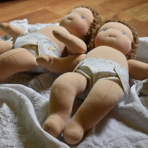 May include: Two cloth dolls with brown hair and blue eyes are lying on a white crocheted blanket. They are wearing white and yellow patterned cloth diapers.