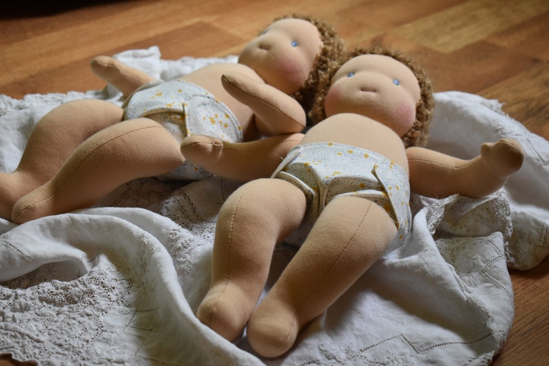 May include: Two cloth dolls with brown hair and blue eyes are lying on a white crocheted blanket. They are wearing white and yellow patterned cloth diapers.