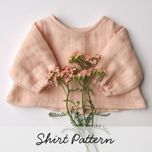 Peut inclure: Un chemisier à manches longues de couleur rose pâle avec un col rond et des poignets froncés. Le chemisier est posé à plat sur une surface blanche avec un bouquet de petites fleurs roses au centre.