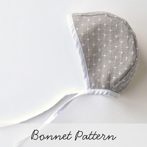 May include: A gray baby bonnet with a white trim and white cross pattern. The bonnet has white ties.