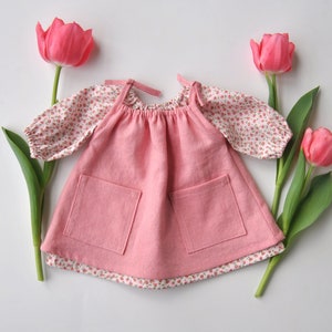 May include: A pink sleeveless dress with two pockets and a floral print top underneath. The dress is surrounded by pink tulips.