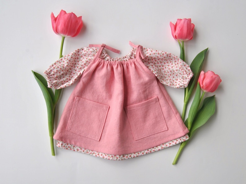 May include: A pink sleeveless dress with two pockets and a floral print top underneath. The dress is surrounded by pink tulips.