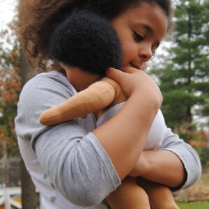 May include: A young girl with curly brown hair is holding a brown doll with black hair. She is wearing a grey and white polka dot shirt.