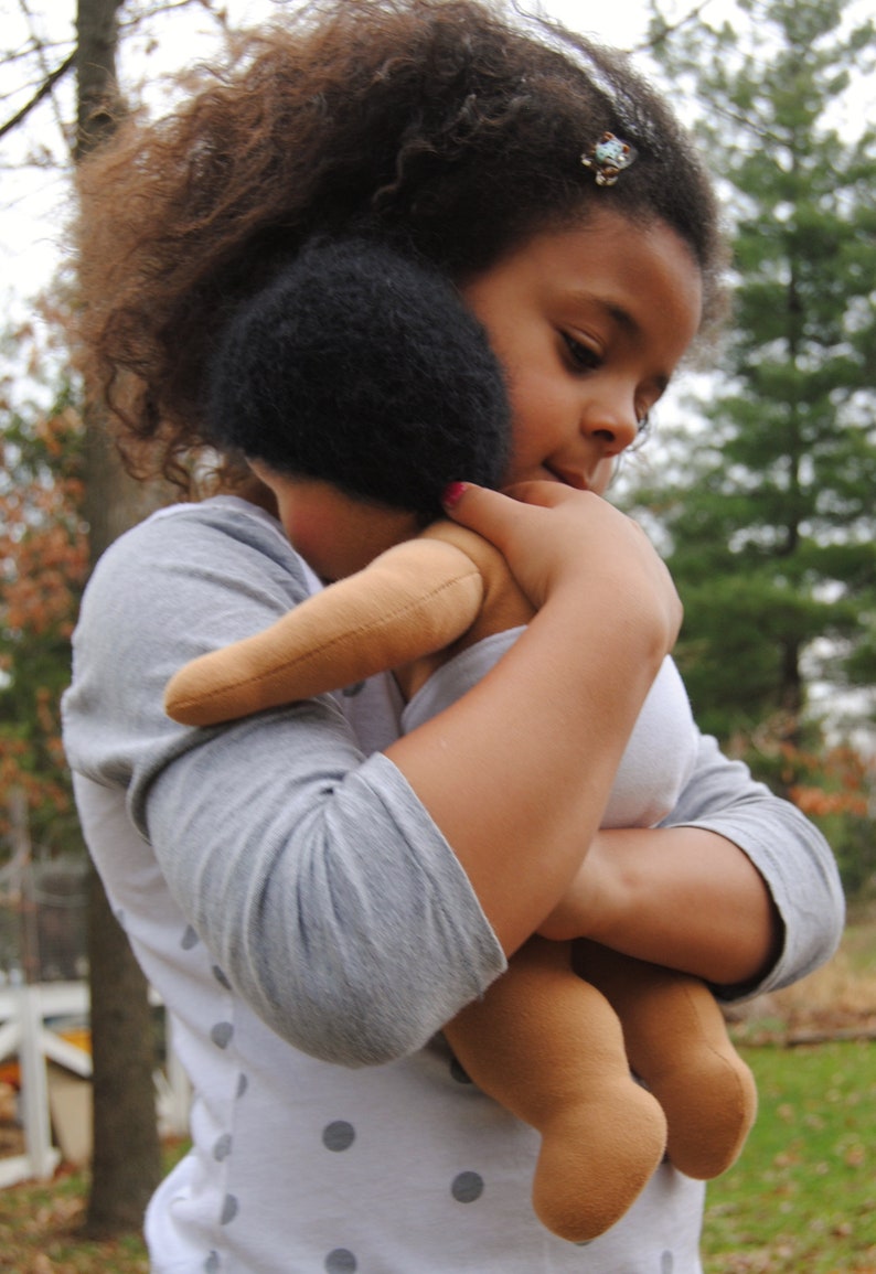 May include: A young girl with curly brown hair is holding a brown doll with black hair. She is wearing a grey and white polka dot shirt.
