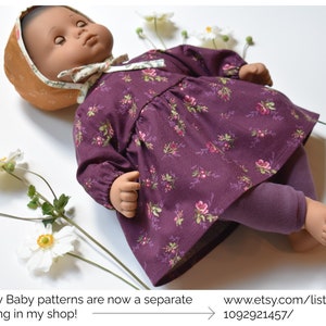 May include: A doll wearing a purple floral dress with long sleeves and purple leggings. The doll is wearing a brown and white hat. The doll is lying on a white surface with white flowers.