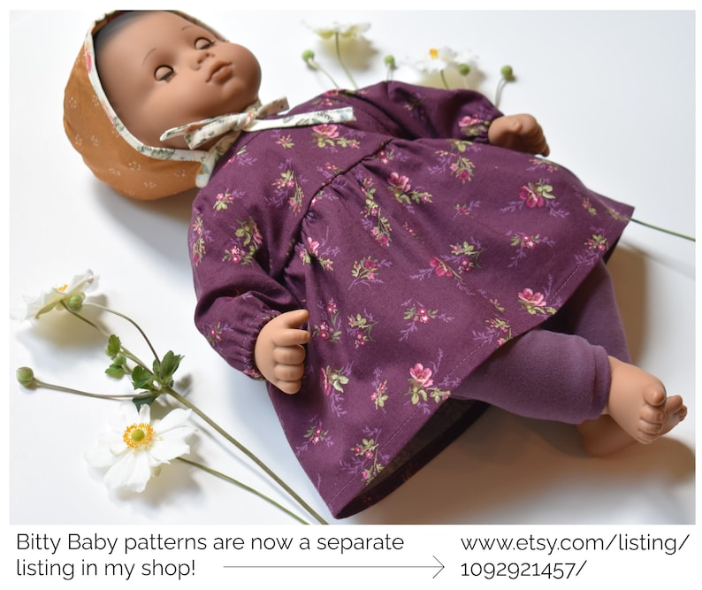 May include: A doll wearing a purple floral dress with long sleeves and purple leggings. The doll is wearing a brown and white hat. The doll is lying on a white surface with white flowers.
