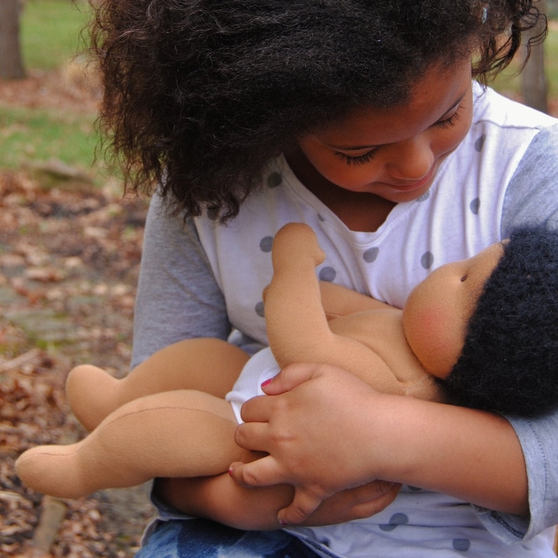 May include: A young girl with dark curly hair is holding a brown cloth doll with black hair. The doll is wearing a white diaper and has a rosy complexion.