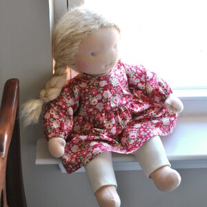 May include: A handmade doll with blonde hair, wearing a red floral dress and white pants. The doll is sitting on a windowsill.