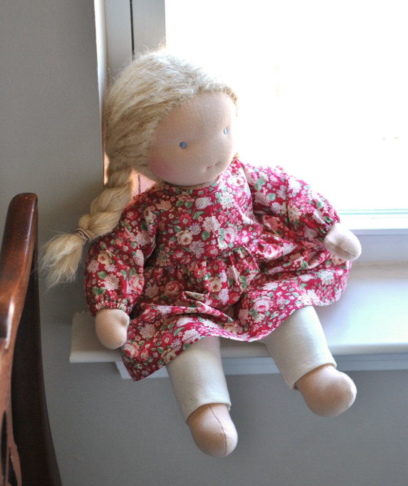 May include: A handmade doll with blonde hair, wearing a red floral dress and white pants. The doll is sitting on a windowsill.