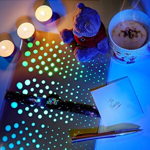 May include: A blue teddy bear sits on a table with a cup of hot chocolate, a note that says "To Santa", a pen, and a glow-in-the-dark sticker sheet with a galaxy design. The sticker sheet has a label that says "Star Scenes".