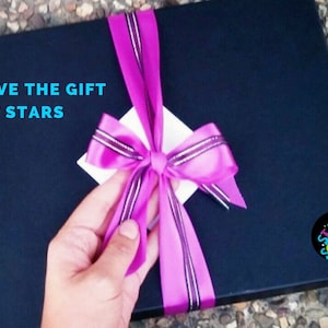 May include: A black gift box with a pink ribbon and bow. The box has a white label with the text "GIVE THE GIFT OF STARS" and an arrow pointing to the right. The box also has a round sticker with the text "STAR SCENES" in colorful letters.