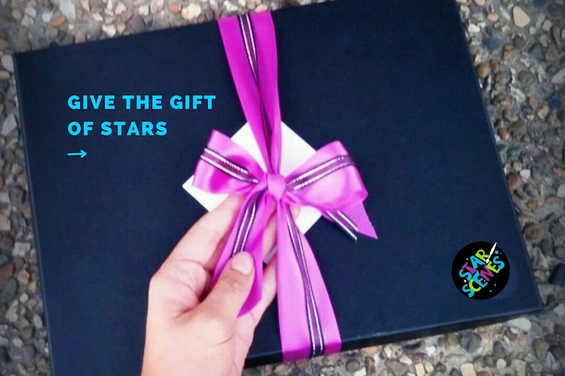 May include: A black gift box with a pink ribbon and bow. The box has a white label with the text "GIVE THE GIFT OF STARS" and an arrow pointing to the right. The box also has a round sticker with the text "STAR SCENES" in colorful letters.