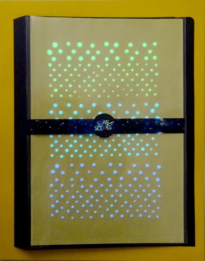 Puede incluir: Una libreta amarilla con una cubierta negra y una banda negra con el texto "STAR SCENES" en blanco. La libreta tiene un patr&oacute;n de peque&ntilde;os puntos blancos, azules y verdes que brillan en la oscuridad.