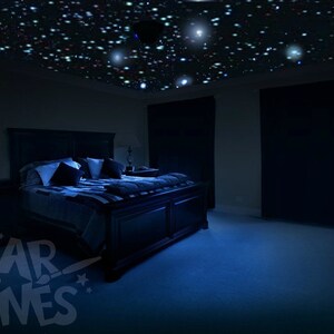 Puede incluir: Un dormitorio oscuro con una cama hecha con almohadas blancas y negras y una manta azul. El techo est&aacute; cubierto con un efecto de cielo estrellado.