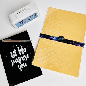 May include: A yellow craft stencil with a pattern of small circles in a gradient design. The stencil is packaged in a clear plastic sleeve and has a black and blue sticker with the text "STAR SCEIVES" on it. The stencil is on a white surface with a black notebook and a white clock.