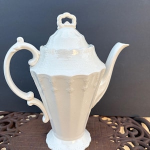 May include: A white porcelain teapot featuring a faceted body with vertical panels, a curved handle, a pointed spout, and a matching lid with a decorative finial. The surface has a subtle embossed texture and a glossy finish throughout the piece.