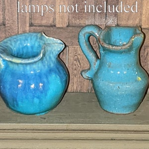 Puede incluir: Dos pequeñas jarras de cerámica azul con un aspecto rústico y hecho a mano. Las jarras están en un estante de madera con el texto "lámparas no incluidas" encima.
