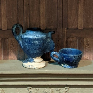 May include: A blue and white ceramic teapot and a matching cup. The teapot has a handle and a spout. The cup has a handle. Both pieces are glazed in a blue color with a white base.