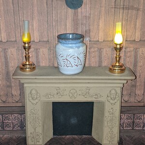 Peut inclure: Un manteau de cheminée miniature avec un pot en céramique et deux lampes en laiton. Le pot est blanc avec un bord bleu et un motif de poisson répété. Les lampes ont une lumière jaune et sont de chaque côté du pot.