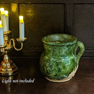 May include: A green glazed ceramic jug with a handle. The jug has a rustic, handmade look and is sitting on a wooden surface.