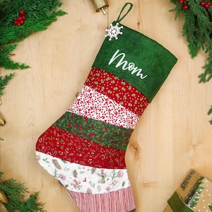 Op de afbeelding: Een kerstkous met een donkergroene boord met de witte geborduurde tekst Mom en een wit sneeuwvlokornament. De kous bestaat uit horizontale stroken stof in rode, witte en groene patronen, waaronder bessen, dennenappels en strepen.