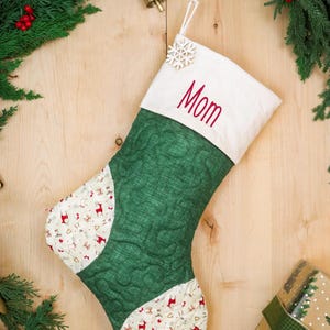 Könnte beinhalten: Ein gesteppter Weihnachtsstrumpf mit einem dunkelgrünen Hauptteil und einer weißen Manschette, die mit dem Wort Mom in rotem Garn bestickt ist. Die Zehen- und Fersenbereiche sind weiß mit einem Muster aus roten Rentieren und kleinen goldenen Sternen.
