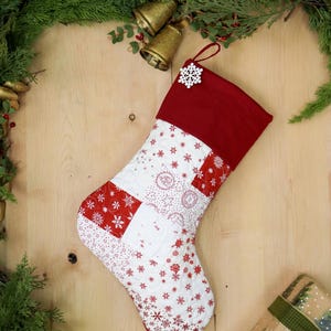 Op de afbeelding: Een gewatteerde kerstsok met een rode manchet en een sneeuwvlokkenpatroon. De sok heeft een patchwork-ontwerp met rode en witte stoffen. Een wit sneeuwvlokornament is bevestigd. De afbeelding bevat groen en gouden bellen.
