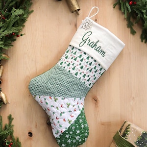 以下が含まれることがあります： 緑色で「Graham」と刺繍された、キルティング加工のクリスマスストッキング。ツリー、雪の結晶、ギンガムチェックなど、緑と白の模様のパッチワークデザインです。雪の結晶の飾りと吊り下げ用のループが上部にあります。