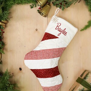 Peut inclure: Une chaussette de Noël avec le nom "Baylee" brodé en rouge sur le dessus blanc. La chaussette présente des sections de tissu blanc matelassé et de tissu rouge à motifs. Un flocon de neige est près du haut.