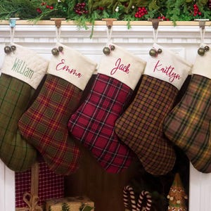 Puede incluir: Cinco calcetines navideños de tela colgados en fila, cada uno con un patrón de cuadros único en tonos verde, rojo y marrón. Cada calcetín tiene un puño de color crema bordado con los nombres William, Emma, Jack y Kaitlyn.