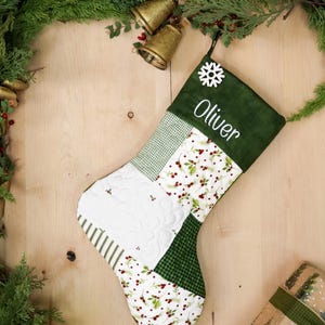 以下が含まれることがあります： 雪の結晶と「オリバー」という名前が刺繍された、緑と白のパッチワークのクリスマスストッキング。ストッキングは、赤いベリーとヒイラギの葉の模様で飾られています。
