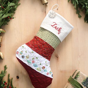 以下が含まれることがあります： 赤い文字で「Zach」と刺繍された白いカフが付いた、キルティングのクリスマスストッキング。ストッキングは、赤い模様の生地、緑と白のチェック柄、カラフルなオーナメントデザインの白い部分で構成されています。上部には雪の結晶のチャームが付いています。