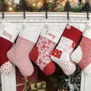 Peut inclure: Cinq bas de Noël rouges et blancs sont suspendus à une cheminée. Chaque bas présente un motif différent, notamment des rayures, des imprimés floraux et des motifs matelassés. Le haut de chaque bas est blanc, et certains sont personnalisés avec des noms.