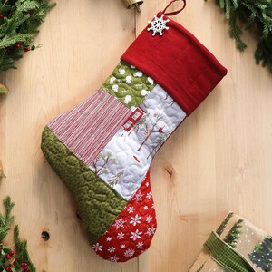 以下が含まれることがあります： 赤いカフと雪の結晶のアクセントが付いた、キルト加工のクリスマスストッキング。パッチワークのデザインには、赤と白のストライプ、緑のテクスチャード加工生地、赤い家のある冬のシーンが含まれています。緑の木と緑のリボンが付いた茶色の紙に包まれたギフトも表示されます。