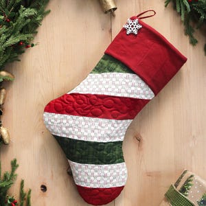 Peut inclure: Une chaussette de Noël matelassée avec des sections rayées rouges, vertes et blanches. Le haut et le bout sont rouges, avec un charme de flocon de neige blanc. La chaussette est décorée d'un motif matelassé et est posée sur un fond en bois avec des décorations festives.