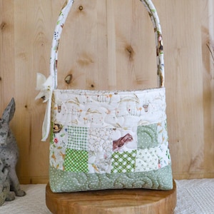 Może przedstawiać: Pikowany kosz wielkanocny z patchworkowym wzorem. Kosz ma białą rączkę z kwiatowym wzorem i białą kokardę. Korpus to mieszanka białych, zielonych i wzorzystych tkanin z motywami królików i kwiatów.