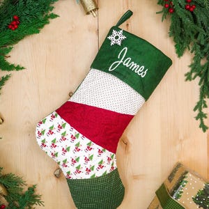 Pode incluir: Uma meia de Natal acolchoada com um punho verde-escuro com o nome James bordado em branco, um detalhe de floco de neve branco e painéis diagonais de tecido com bolinhas brancas, vermelho sólido e uma estampa festiva de Papai Noel.