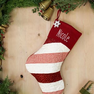 Könnte beinhalten: Roter und weißer gestreifter Weihnachtsstrumpf mit Karomuster und weißen Punkten. Der Strumpf hat einen roten Bund mit dem Namen "Nicole" bestickt und eine weiße Schneeflocke als Verzierung.