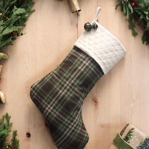 May include: A Christmas stocking with a green and brown plaid pattern, topped with a quilted white cuff. The stocking is decorated with small bells and is surrounded by festive greenery and a wrapped gift.