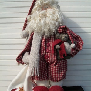 May include: A handmade Santa Claus doll in a red and white checked outfit, long hat, and holding a small teddy bear. The doll has a white beard, red shoes, and a white blanket with star and moon appliqués. A festive Christmas decoration.