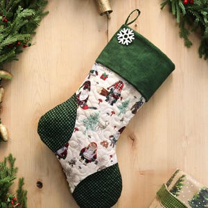 以下が含まれることがあります： 緑色の袖口とヒール、白い雪の結晶ボタンが付いた、キルト加工のクリスマスストッキング。メインボディには、ノーム、木、キノコのパターンがあります。ストッキングは緑と白のチェック柄の生地で飾られています。