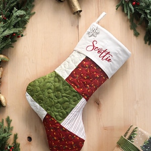 以下が含まれることがあります： 「Scottie」という名前が赤い筆記体で刺繍された白い袖口が付いた、キルト加工のクリスマスストッキング。ストッキングは、緑、赤の花柄、白の模様の生地のパッチワークセクションと、雪の結晶のチャームが特徴です。