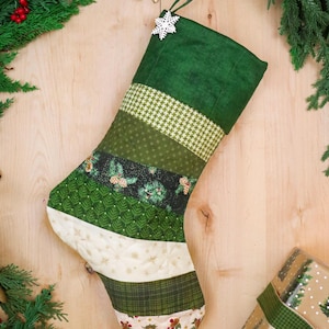 May include: A quilted Christmas stocking featuring horizontal fabric strips in shades of green, cream, and white. The design includes patterns of houndstooth, pine trees, pinecones, and holly berries, finished with a white snowflake ornament.