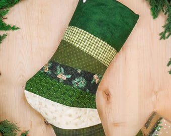 Pode incluir: Uma meia de Natal acolchoada com tiras de tecido horizontais em tons de verde, creme e branco. O design inclui padrões de pé-de-galinha, pinheiros, pinhas e bagas de azevinho, finalizada com um ornamento de floco de neve branco.